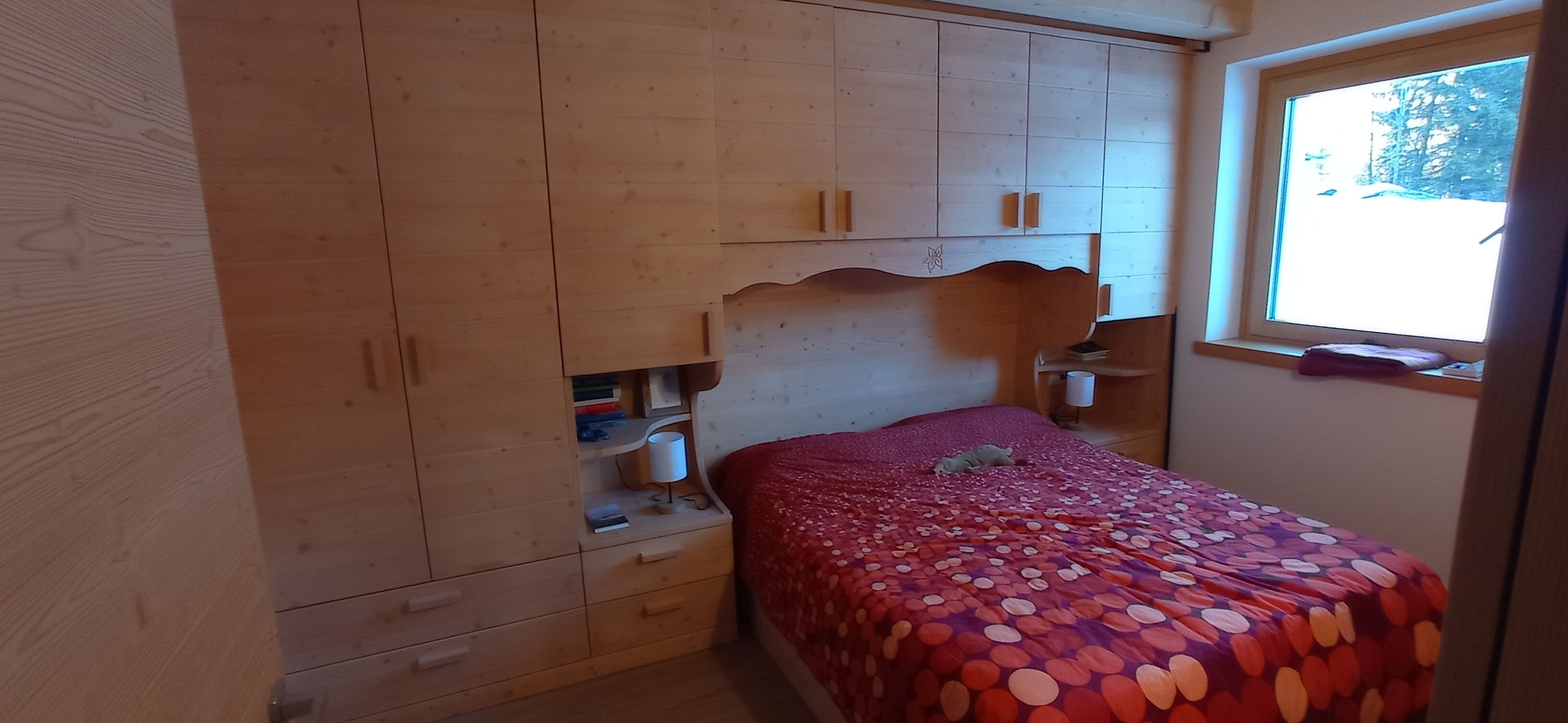 A bedroom with a wooden headboard, a bed with a polka dot bedspread, a nightstand with a lamp, and a window with a view of trees.