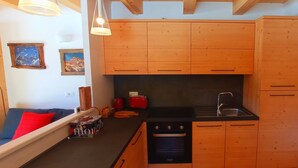 Een kleine keuken met houten kastjes, een zwart aanrecht en een fornuis. Er is een raam met uitzicht op bergen en een rode stoel.