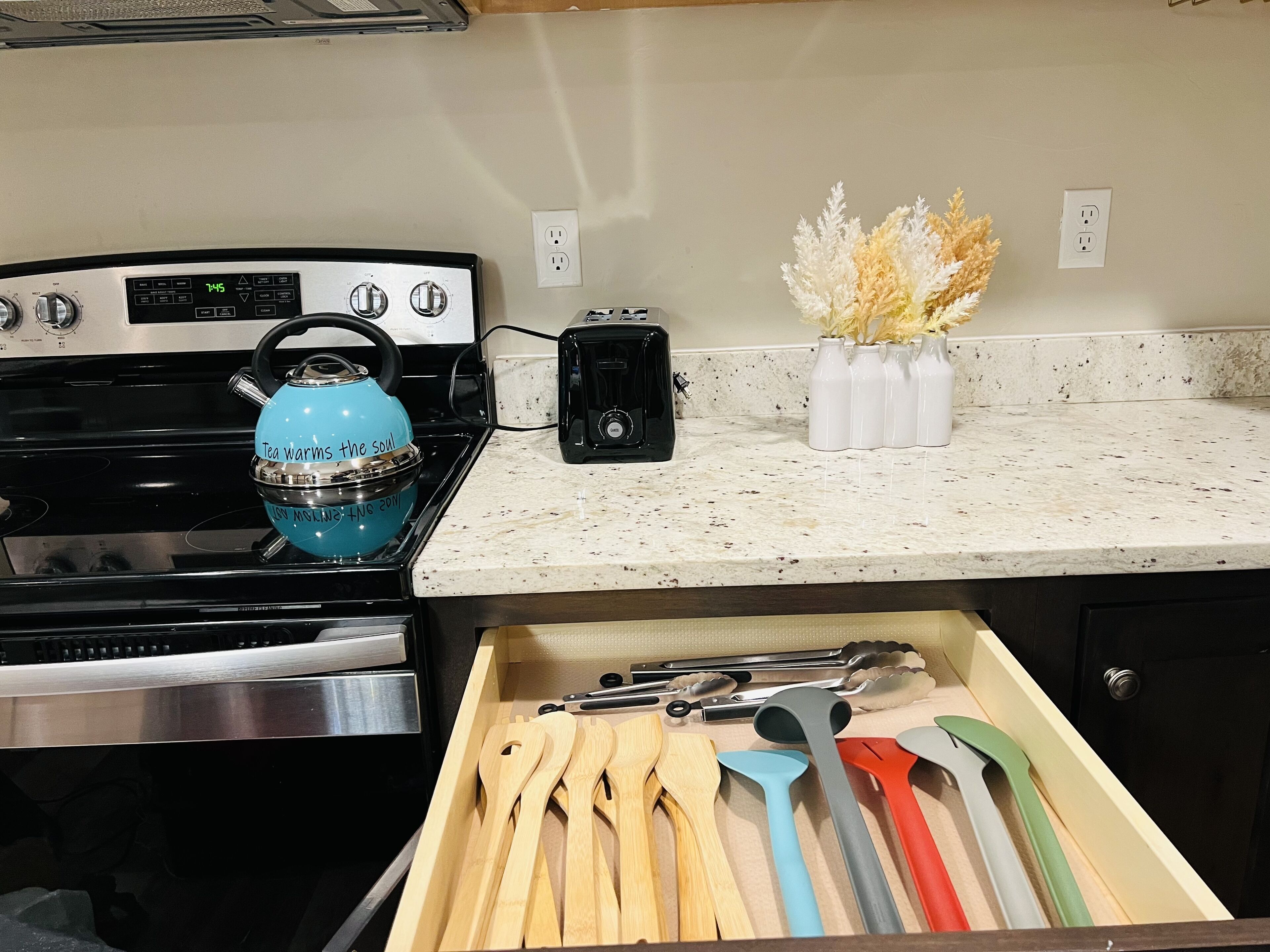 A kitchen with a stove, a blue kettle, a toaster, and a drawer with wooden utensils.