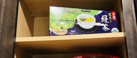 A wooden cabinet with two shelves; the top shelf holds a box of organic green tea, and the bottom shelf contains a vitamin C supplement and a first aid kit.