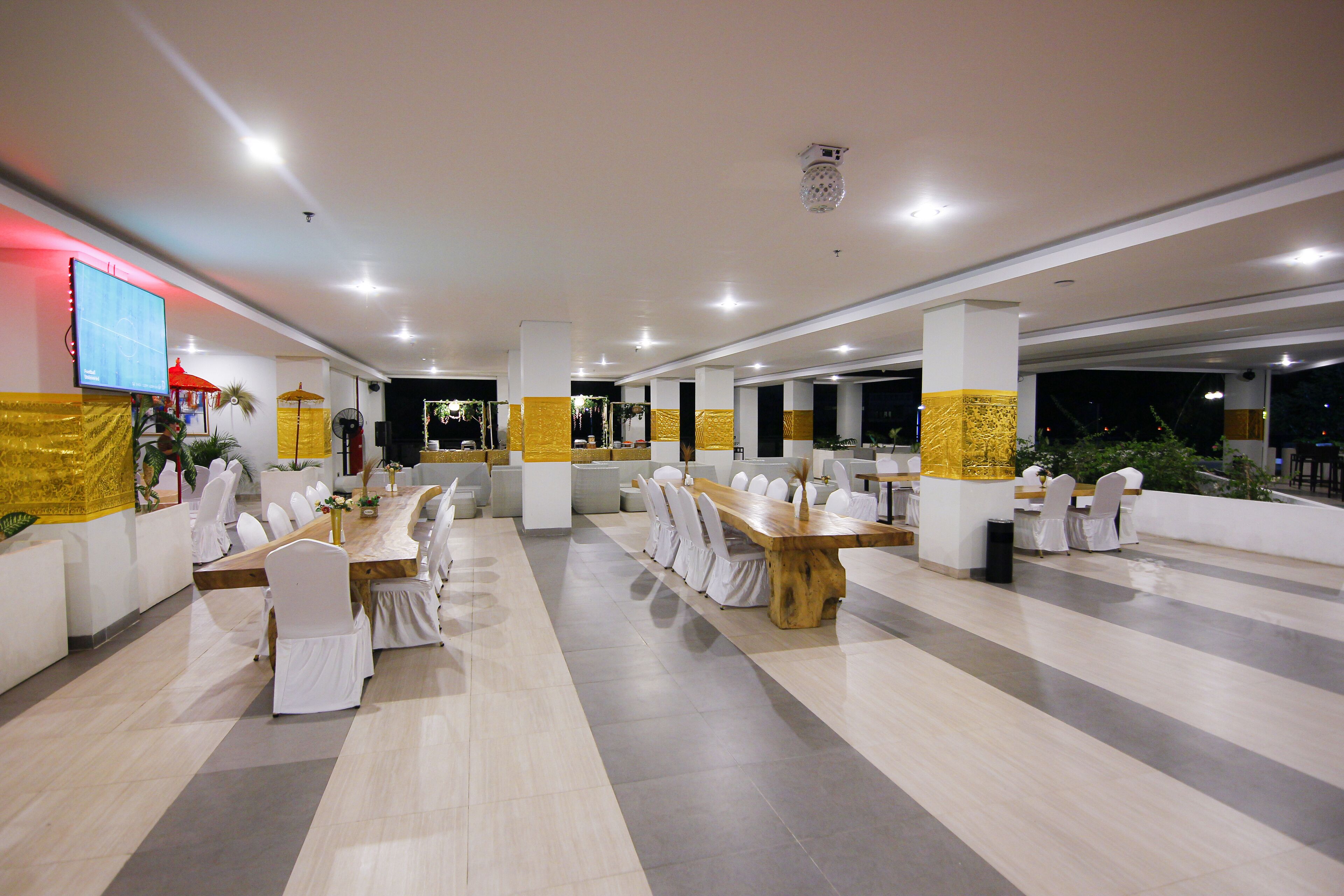 A spacious indoor dining area with multiple tables and chairs, a flat-screen TV mounted on the wall, and a ceiling with recessed lighting.