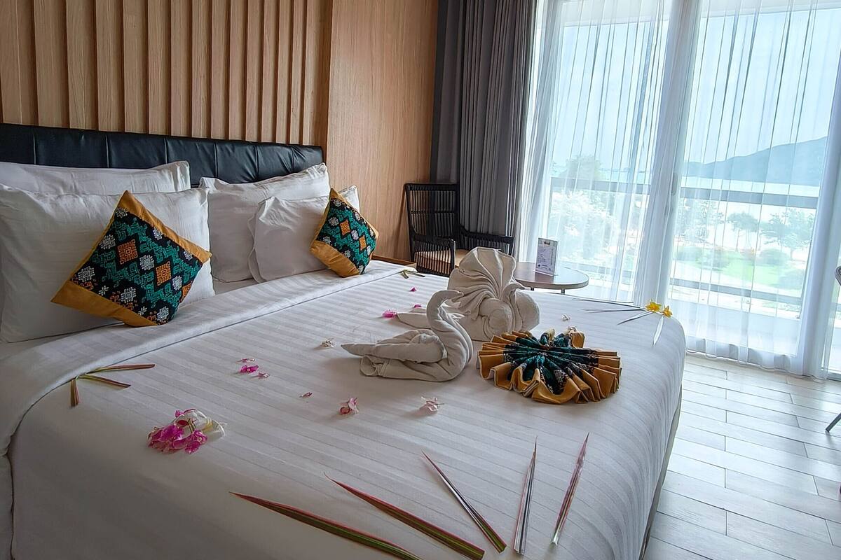 A hotel room with a large bed, wooden panel walls, a view of the outdoors, and a dining area with a table and chairs.