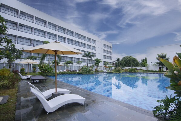 A swimming pool with lounge chairs and umbrellas, surrounded by a modern building with multiple floors and balconies.
