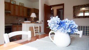 Ein Essbereich mit einem weißen Tisch, Stühlen und einer Vase mit blauen Blumen. Im Hintergrund ist eine Küche zu sehen.