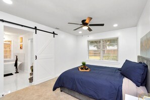 A bedroom with a bed, a ceiling fan, a window with blinds, and a door leading to a bathroom. - Farmhouse Modern Home (Houston)