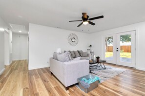 A spacious living room with a grey sofa, a ceiling fan, and a large sliding glass door leading to a backyard. - Farmhouse Modern Home (Houston)