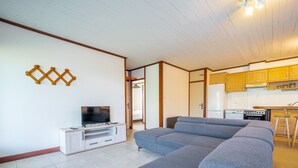 A living room with a grey sofa, a TV on a white entertainment unit, and a kitchen in the background. - Spacious 2-Star House for 10, Near Beach, with Garden and BBQ - Mimizan (Mimizan)