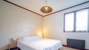 A bedroom with a bed, a nightstand, a wall-mounted lamp, a radiator, and a window with blinds. - Spacious 2-Star House for 10, Near Beach, with Garden and BBQ - Mimizan (Mimizan)
