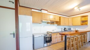 A kitchen with wooden cabinets, a white refrigerator, a stove, a microwave, and a wooden bar stool area. - Spacious 2-Star House for 10, Near Beach, with Garden and BBQ - Mimizan (Mimizan)