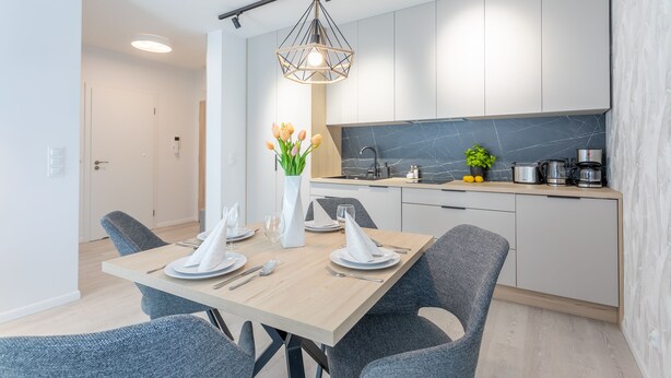 Une cuisine moderne avec des meubles blancs, un pare-feu en marbre et un coin repas comprenant une table en bois et des chaises grises.