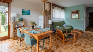 Een gezellige woonkamer met een houten eettafel, stoelen, een bank en een groene fauteuil. Er is een raam met zonwering, een wandlamp en een schilderij aan de muur.