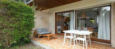 A patio with a wooden table and chairs, a cushioned outdoor seating area, and a view of the interior through glass doors.