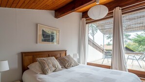 Un dormitorio con techo de madera, una cama con ropa de cama blanca, almohadas a rayas, una mesita de noche con lámpara, un cuadro en la pared y vistas al exterior a través de amplias ventanas.