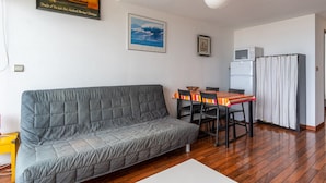 A living room with a sofa, dining table, chairs, a white coffee table, a ceiling light, a refrigerator, and a kitchen area. - Apartment with ocean view, balcony, and all comforts, in Mimizan for 6 people (Mimizan)