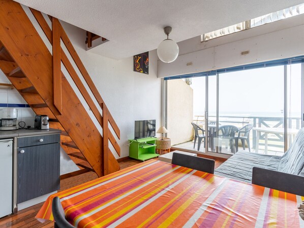 A room with a bed, a desk, a kitchenette, a staircase, and a balcony with a view of the sea.