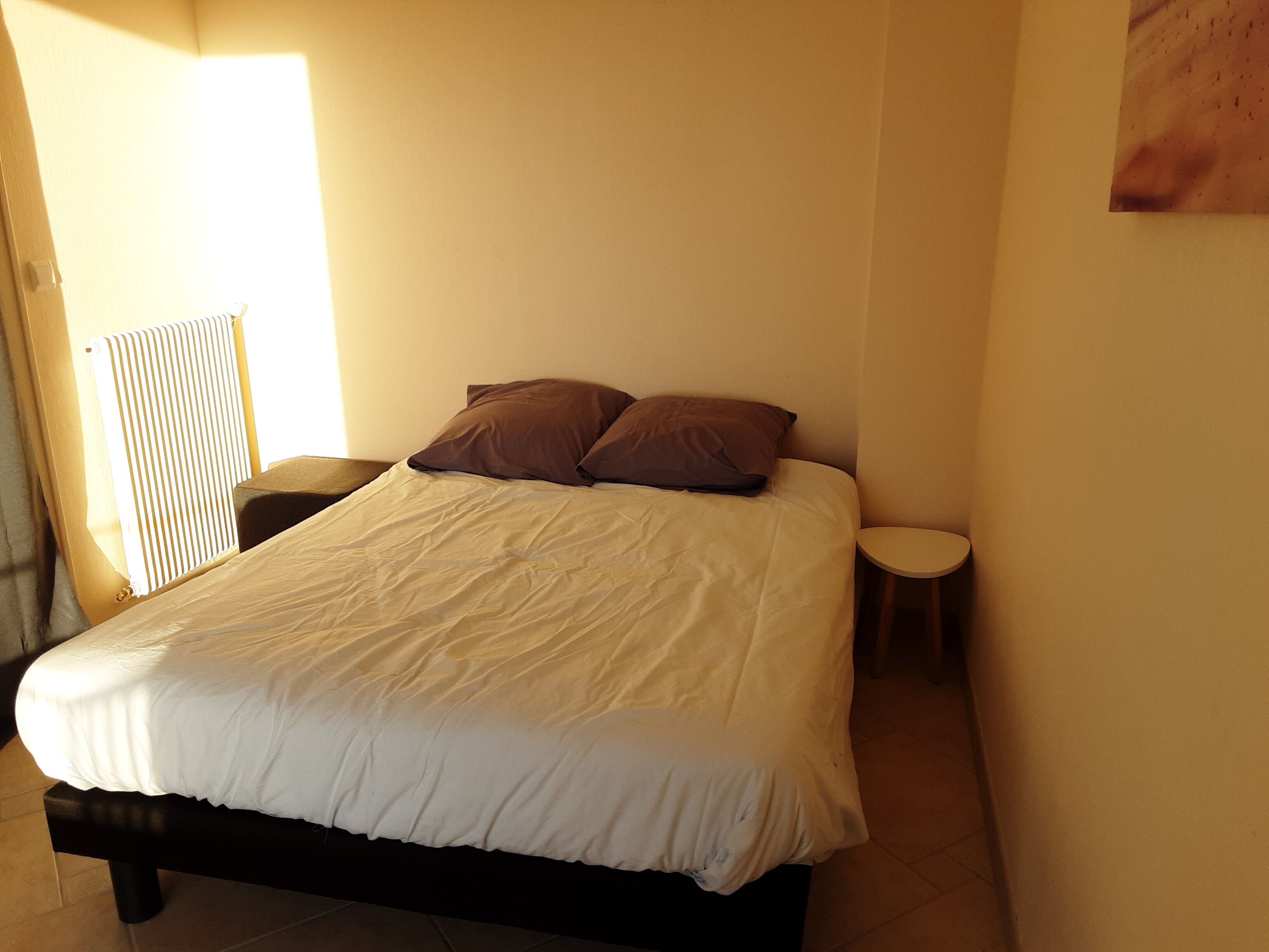 A neatly made bed with a white duvet and two brown pillows in a room with a radiator and a small bedside table.