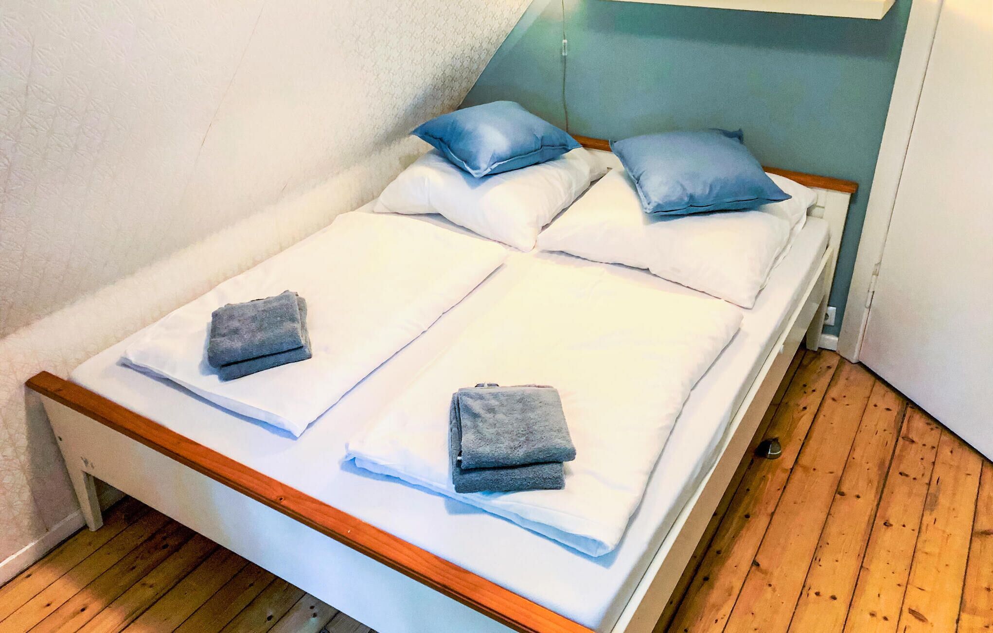 Een eenpersoonsbed met witte beddengoed en twee grijze handdoeken, een houten hoofdbord en een witte muur met een subtiel patroon.