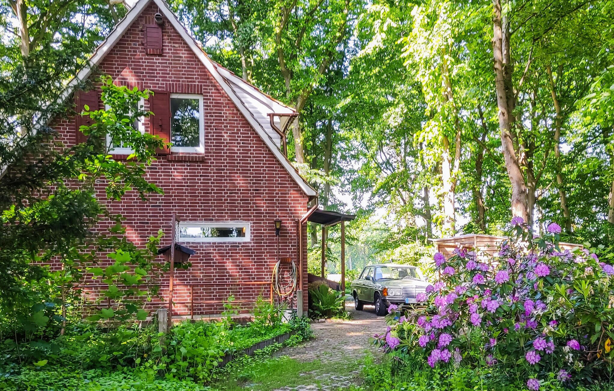 Une maison en briques rouges, avec un toit en pente, entourée d’une végétation luxuriante et de fleurs violettes.