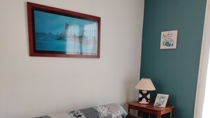 A living room with a patterned sofa, a framed picture of a sailboat, a small table with a lamp, and a wall with a 'Surfing' sign.