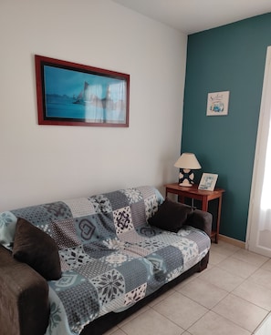 A living room with a patterned sofa, a framed picture of a sailboat, a small table with a lamp, and a wall with a 'Surfing' sign.