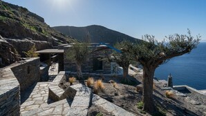 Ein Steinhäuschen mit Terrasse, mit Blick auf eine Küstenlandschaft. Klare Sicht auf das Meer und Hügel im Hintergrund.