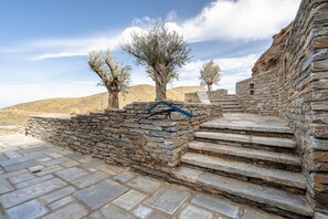 A stone staircase leading up to a stone wall with a blue safety rail, surrounded by olive trees and a clear sky.