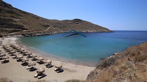 Ein Strand mit Liegestühlen und Sonnenschirmen, ein klares blaues Meer und ein felsiger Hügelhang.
