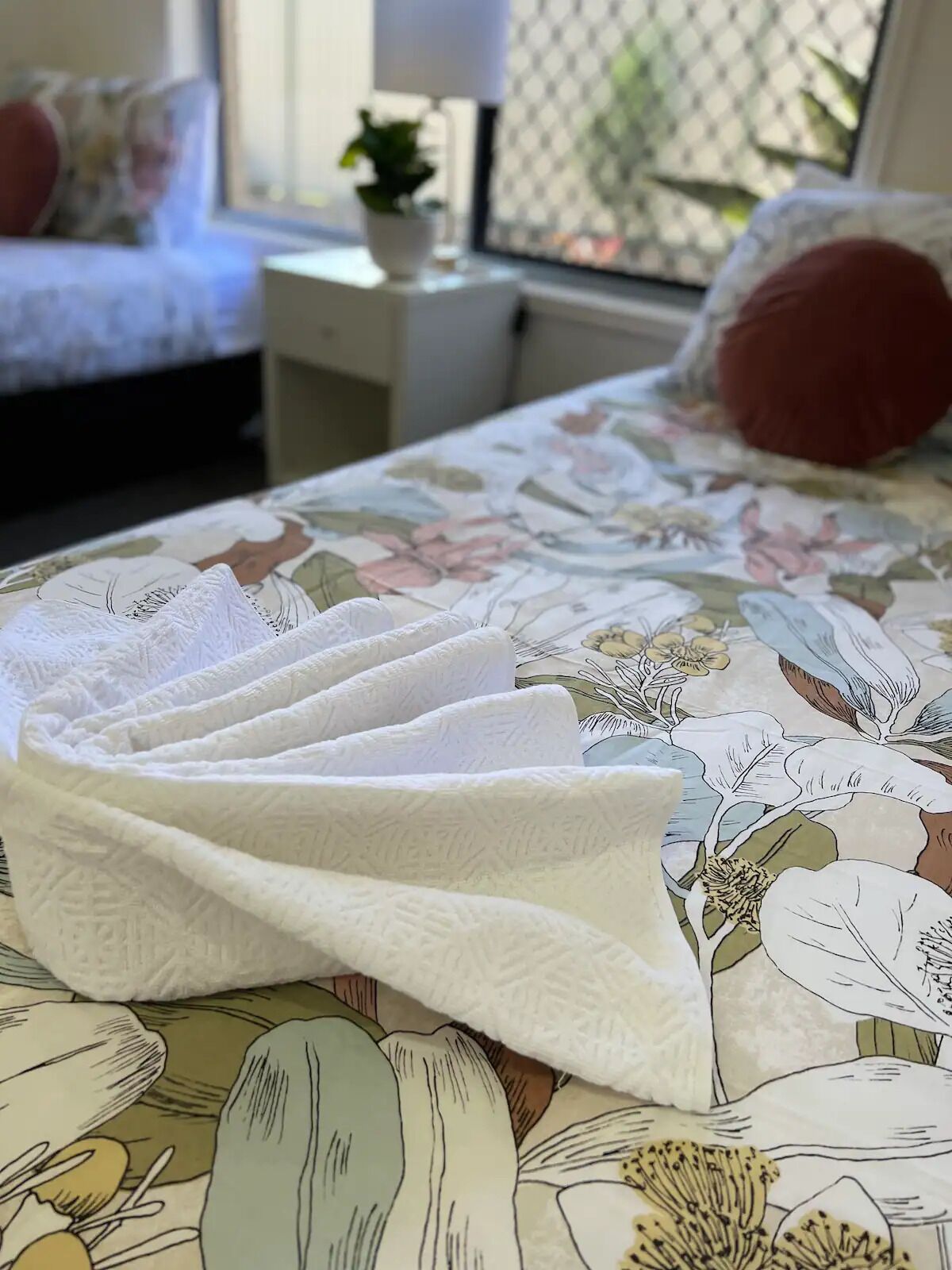Un lit bien fait, recouvert d’une couvre-lit à motifs floraux, avec un oreiller blanc et un oreiller rouge posés sur une table de chevet agrémentée d’une plante.