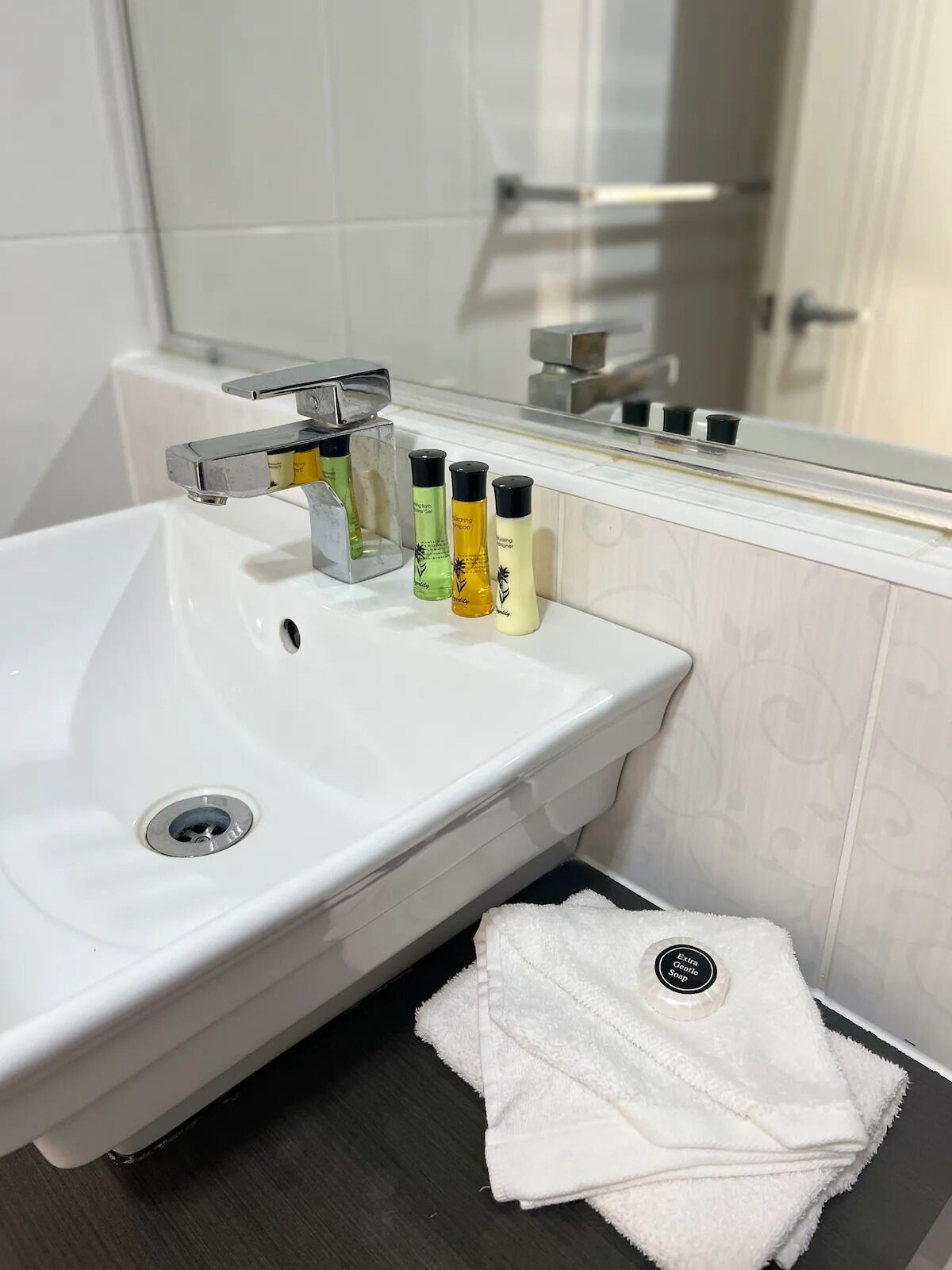 Valkoinen kylpyhuoneen allas, jossa on krominen hana, kolme pulloa shampoota ja valkoinen pyyhe sekä saippua-annostelija.