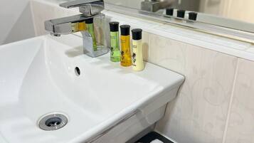 Valkoinen kylpyhuoneen allas, jossa on krominen hana, kolme pulloa shampoota ja valkoinen pyyhe sekä saippua-annostelija.