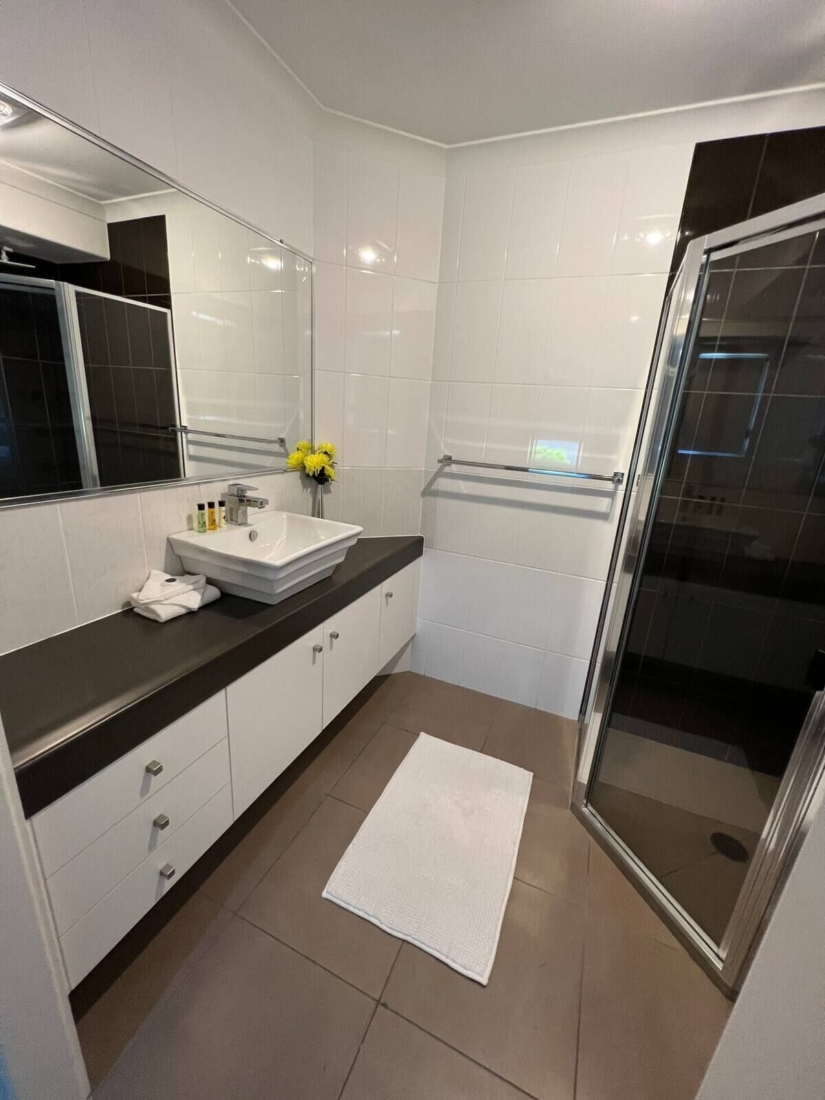 Een moderne badkamer met een grote spiegel, witte tegels, een zwart aanrecht, een witte wastafel en een douchecabine met glazen wanden.