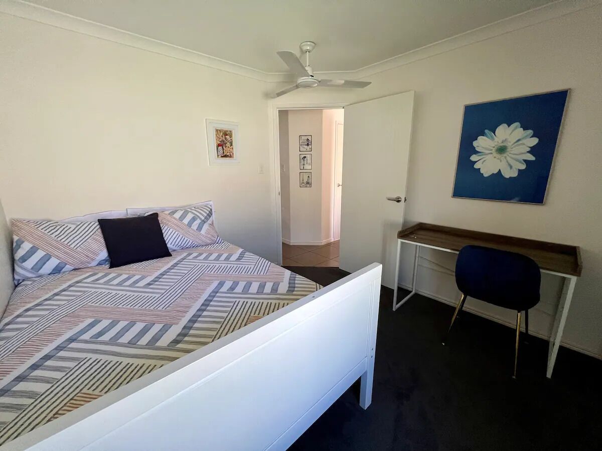 A bedroom with a bed, a desk, a chair, a ceiling fan, and a door leading to another room.