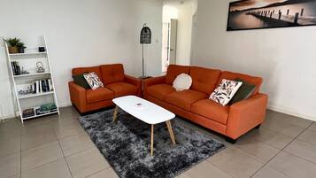 Un salon avec deux canapés orange, une table basse blanche, un tapis à poils longs noir et une bibliothèque blanche.