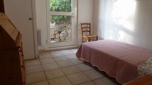 Une pièce comprenant un lit, une chaise, une commode en bois et une fenêtre donnant sur un mur de pierre et la verdure environnante.