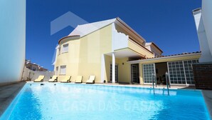 Une piscine avec des chaises longues, un patio couvert et un bâtiment à deux étages surmonté d’un toit en tuiles.