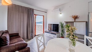 A modern living room with a sofa, a dining table, and a TV. There is a large window with curtains, a plant, and a vase with flowers. - 1 bedroom stunning apartment in Kneze (Kneze)