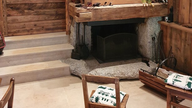 Interior rústico de madera con chimenea de piedra, bancos de madera y una estantería con varios objetos.