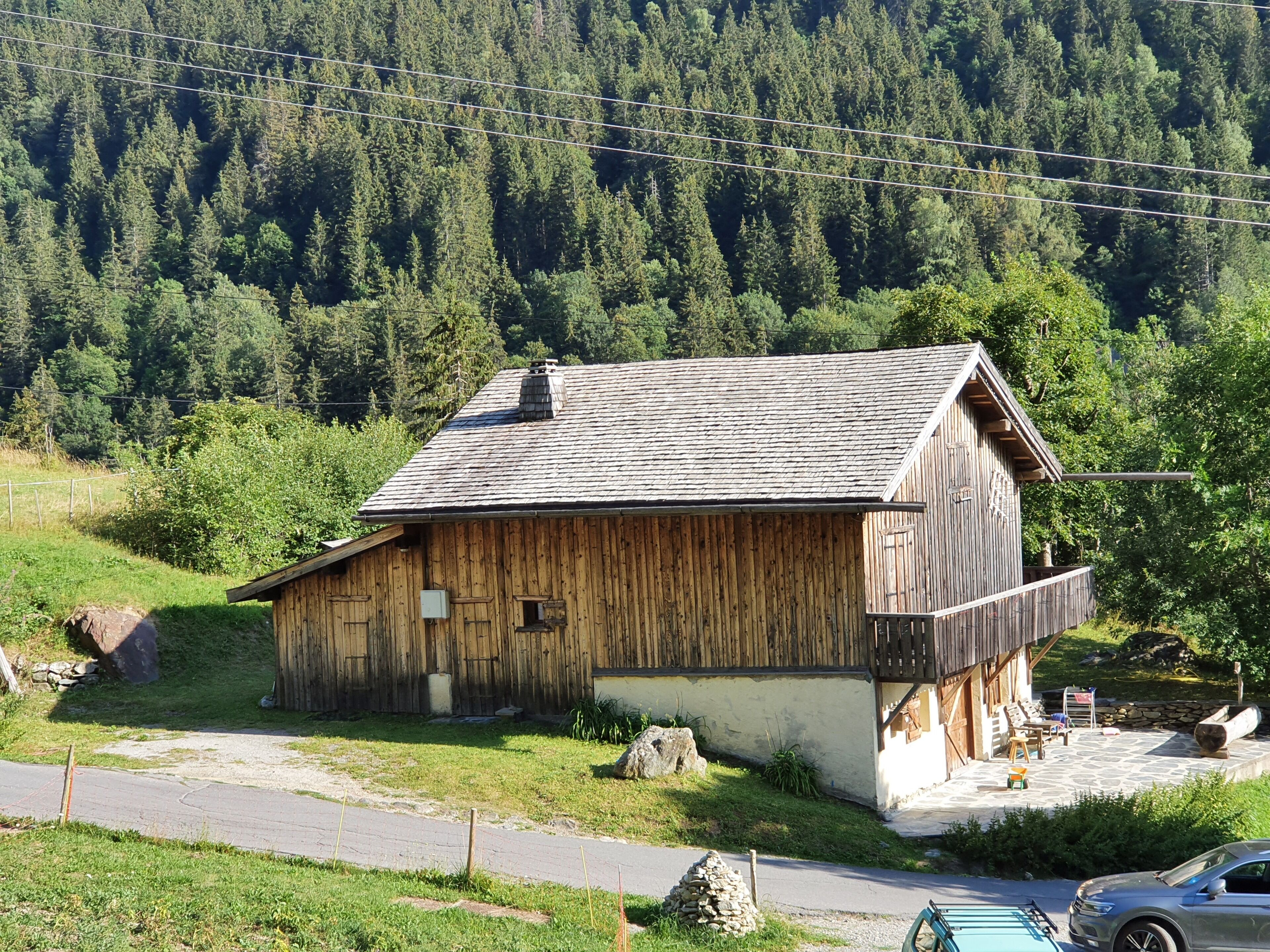 Une cabane en bois rustique, dotée d’un toit en pente, située dans une zone boisée, avec une route goudronnée au premier plan.