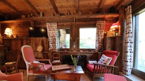 Interior acogedor de una cabaña con paredes de madera, una chimenea y un ventanal con cortinas. Hay dos sillones, una mesa y un sofá.