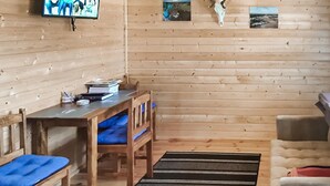 L’intérieur d’un chalet accueillant, avec des murs en bois, un téléviseur fixé au mur, une table en bois avec des livres et une tasse, un banc bleu avec un coussin et un canapé avec un coussin rouge.