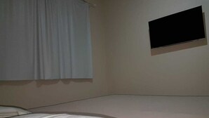 Ein schlichtes Zimmer mit einem Bett, einem Fenster mit Vorhängen und einem an der Wand befestigten Flachbildfernseher.