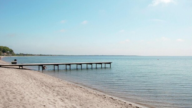 A beach with a wooden pier extending into calm waters, a sandy shore, and a clear sky.
