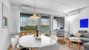A modern living room with a round dining table, white chairs, a grey sofa, and a view of a garden through large windows.