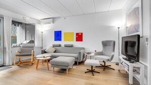 A modern living room with a grey sofa, ottoman, and armchair, a flat-screen TV, and a coffee table. There are three colorful paintings on the wall.