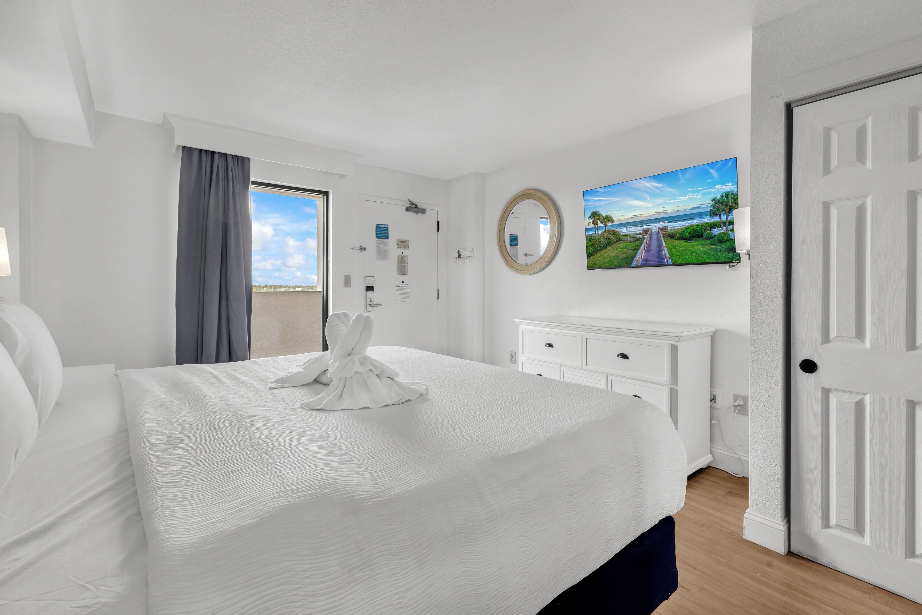 A hotel room with a large bed, a television, a mirror, and a view of the outdoors.