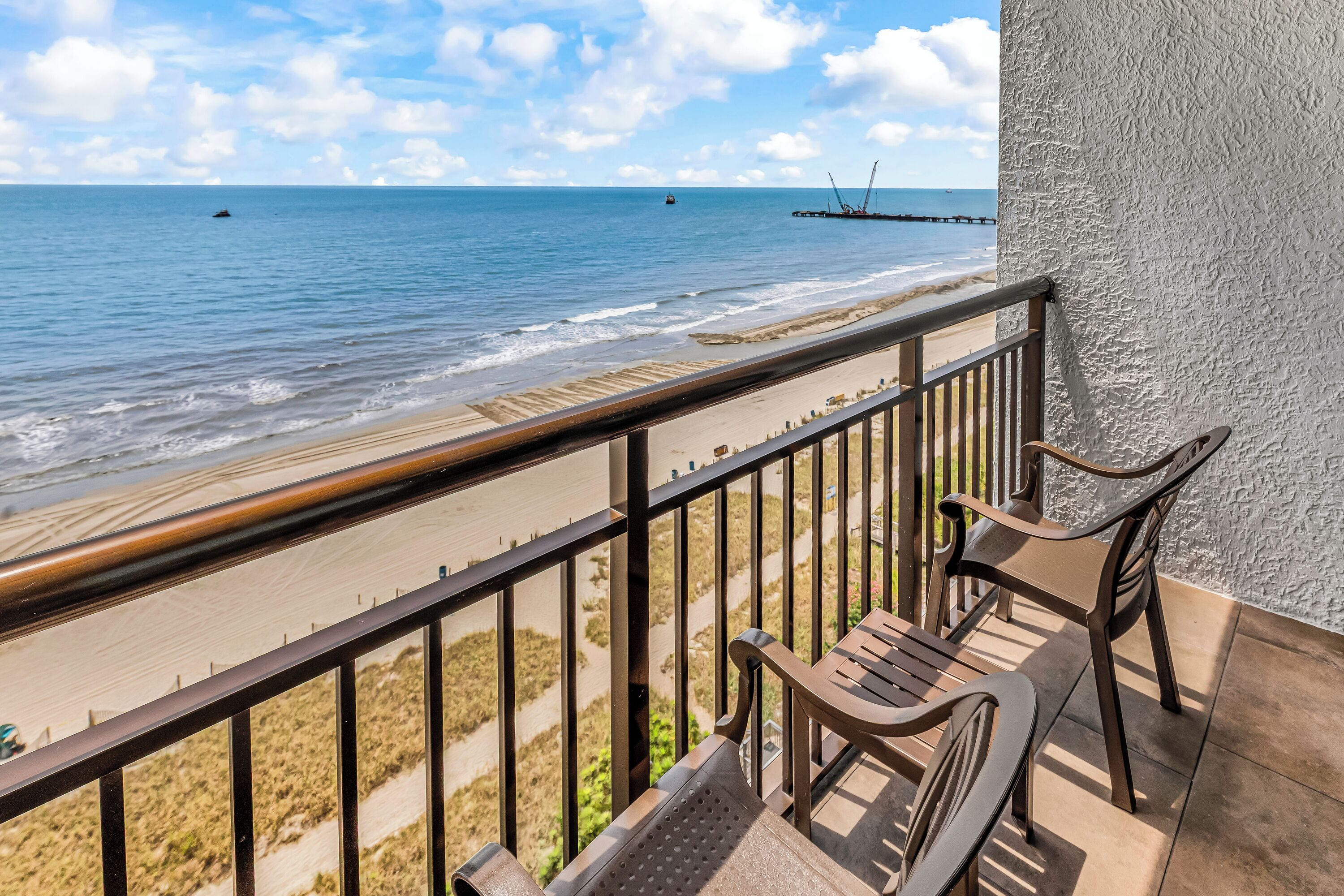 A balcony with a view of the beach, a railing, and two chairs.