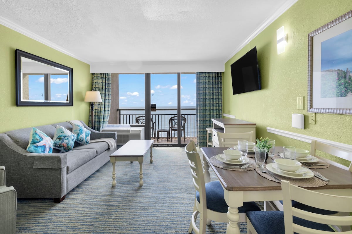 Ein Zimmer mit einer Couch, einem Esstisch, Stühlen, einem Fernseher und einem Balkon mit Blick.