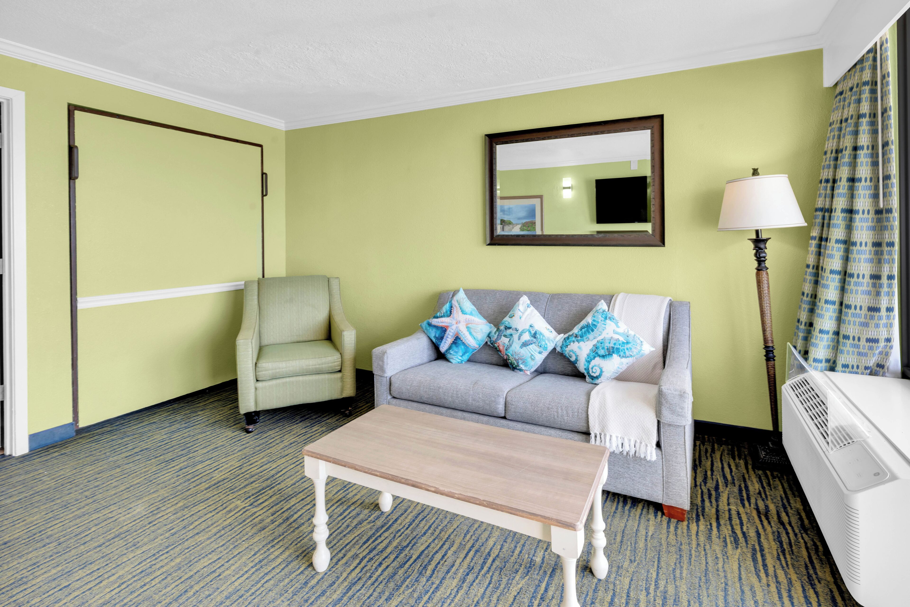 A room with a grey sofa, a small table, a chair, a lamp, and a mirror on the wall.