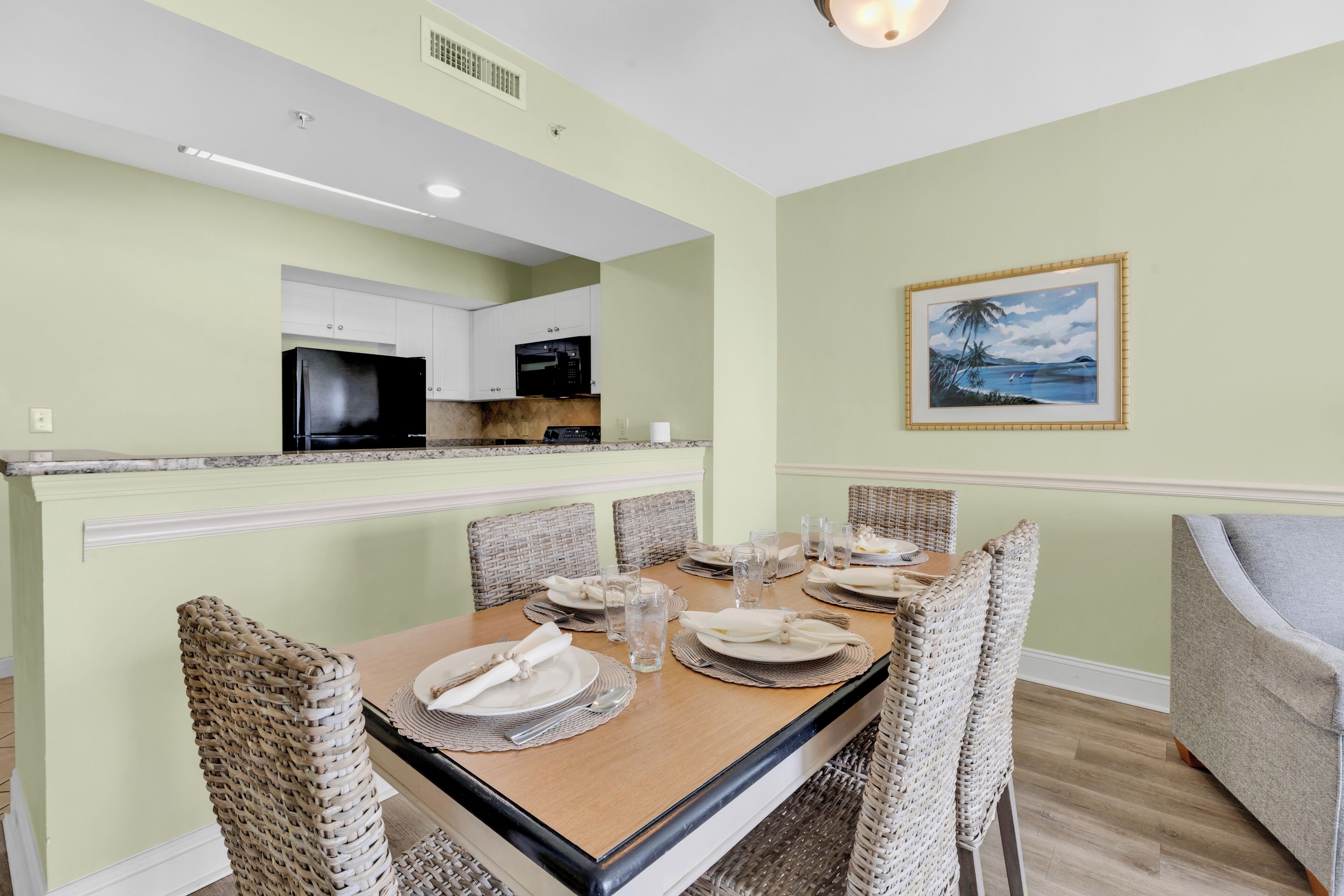 Un coin repas avec une table dressée pour quatre personnes, entourée de chaises en osier, une cuisine en arrière-plan et un tableau encadré au mur.