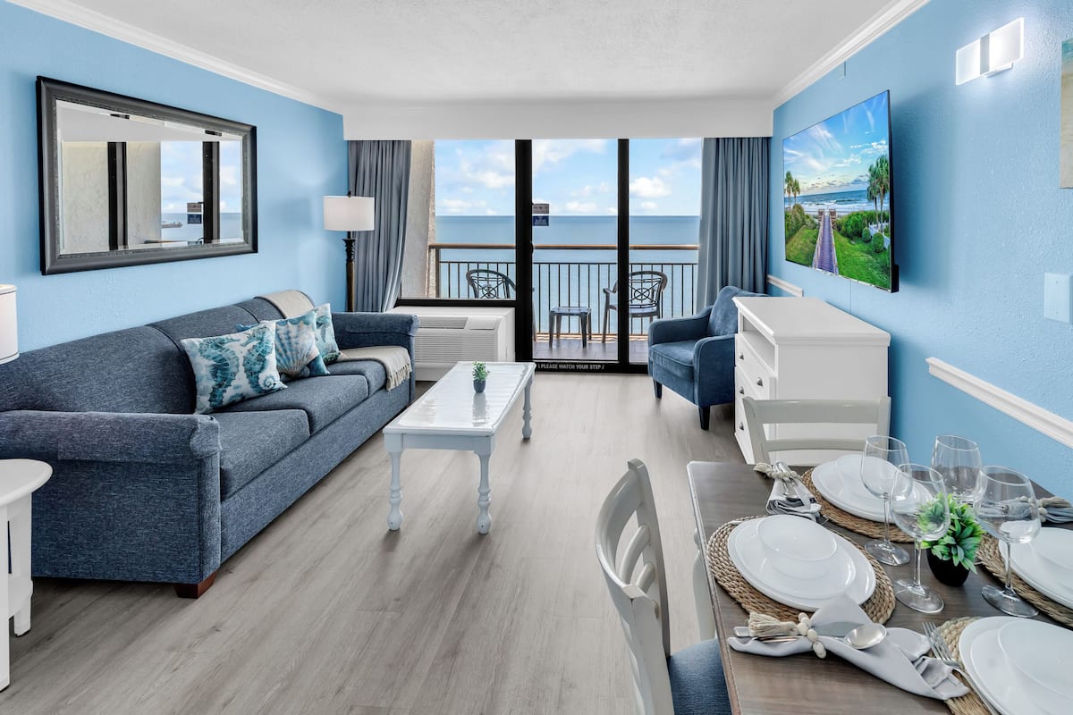 Ein modernes Wohnzimmer mit Sofa, einem Esstisch für vier Personen, einem an der Wand befestigten Fernseher und einem Balkon mit Meerblick.